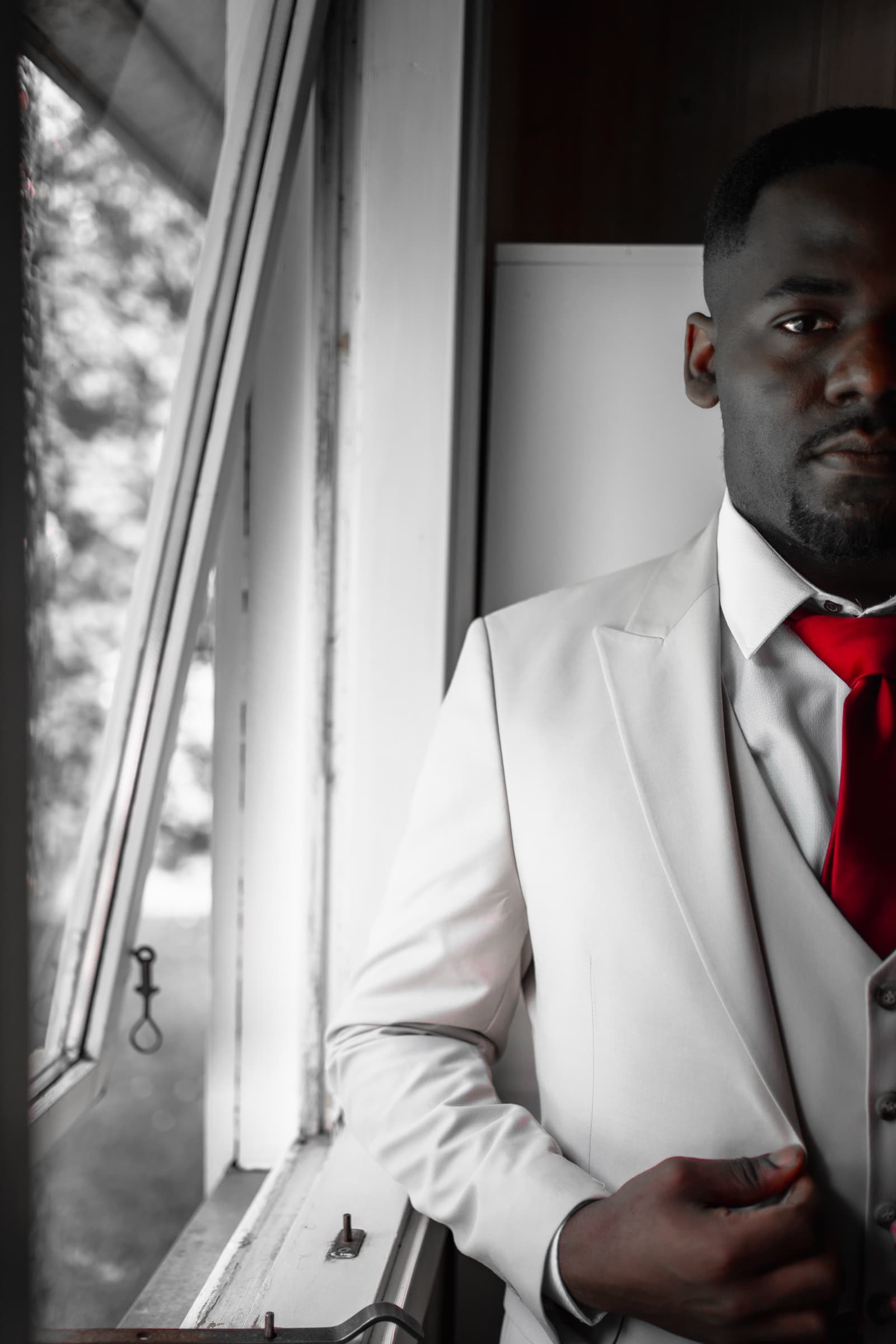 Groom in white three-piece suit and crimson tie, selective-colour portrait by a window