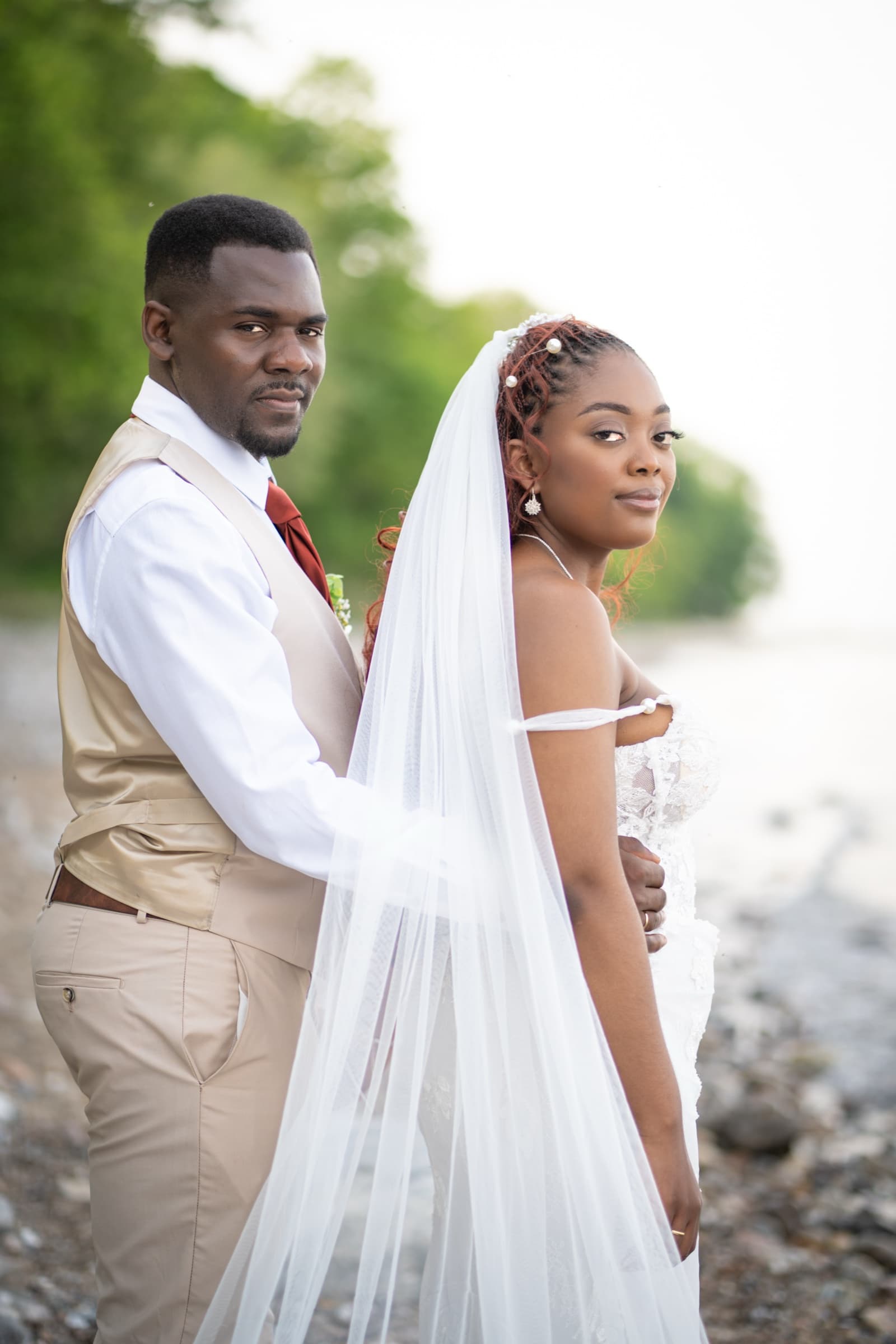 Bride and groom on a pebble shore, the veil trailing across the frame at golden hour