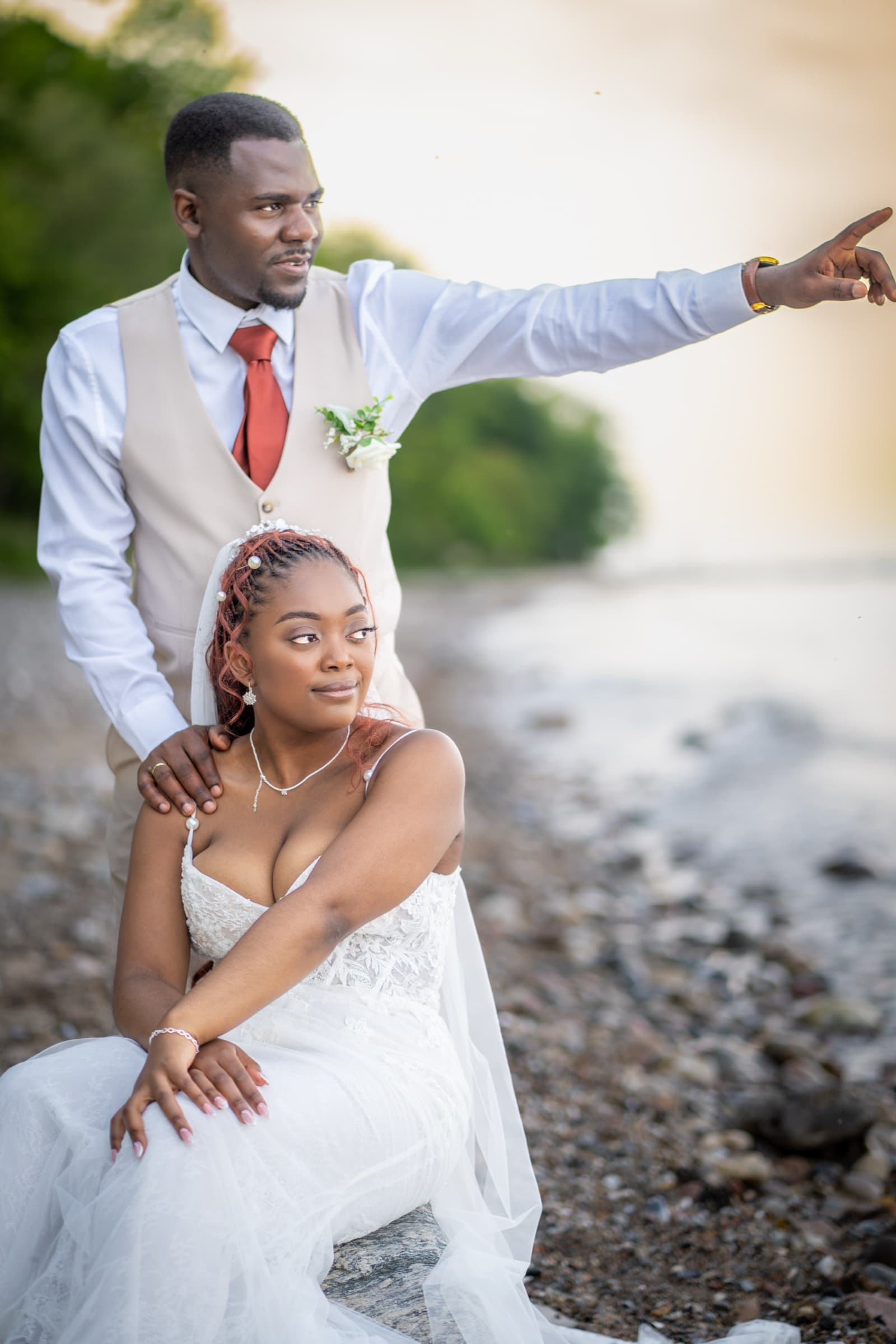 Groom pointing toward the horizon while the bride sits at his side, lakeside sunset