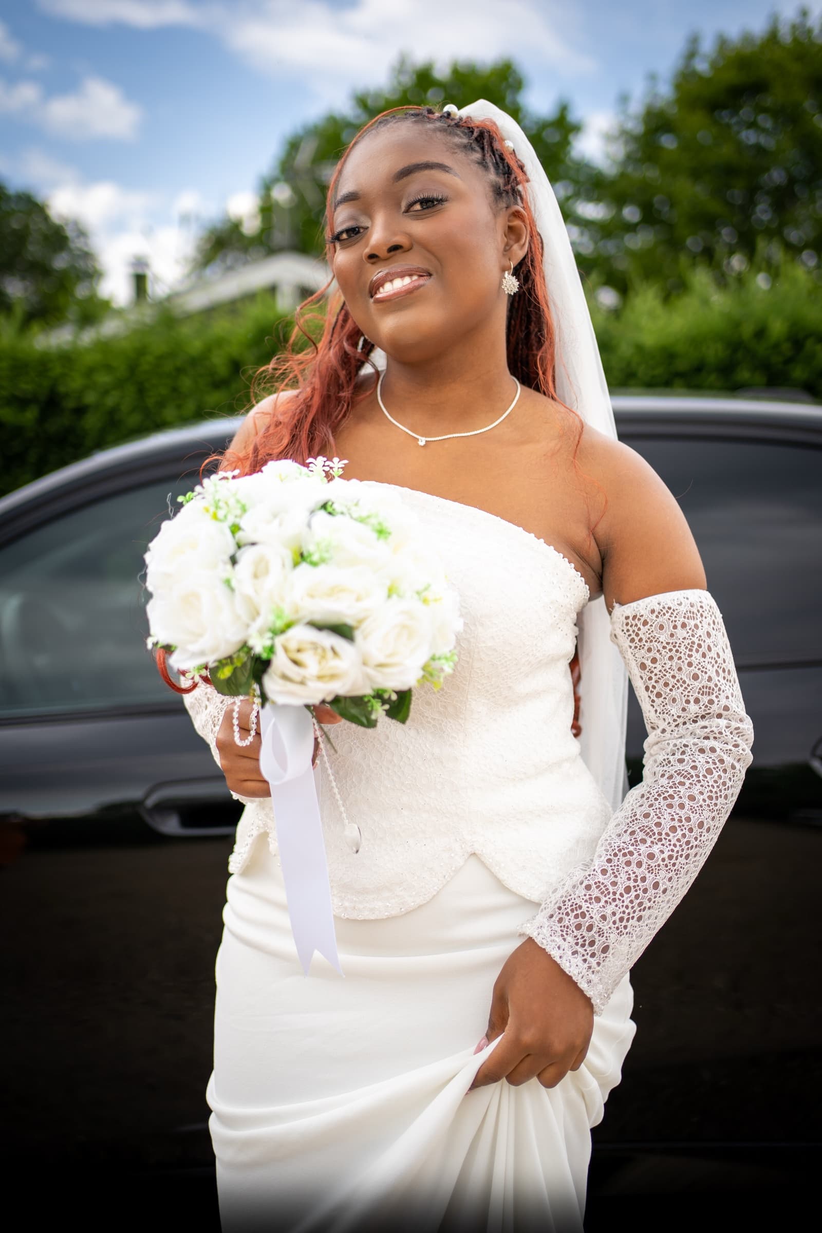 Bride smiling with her bouquet, black wedding car behind her, summer afternoon