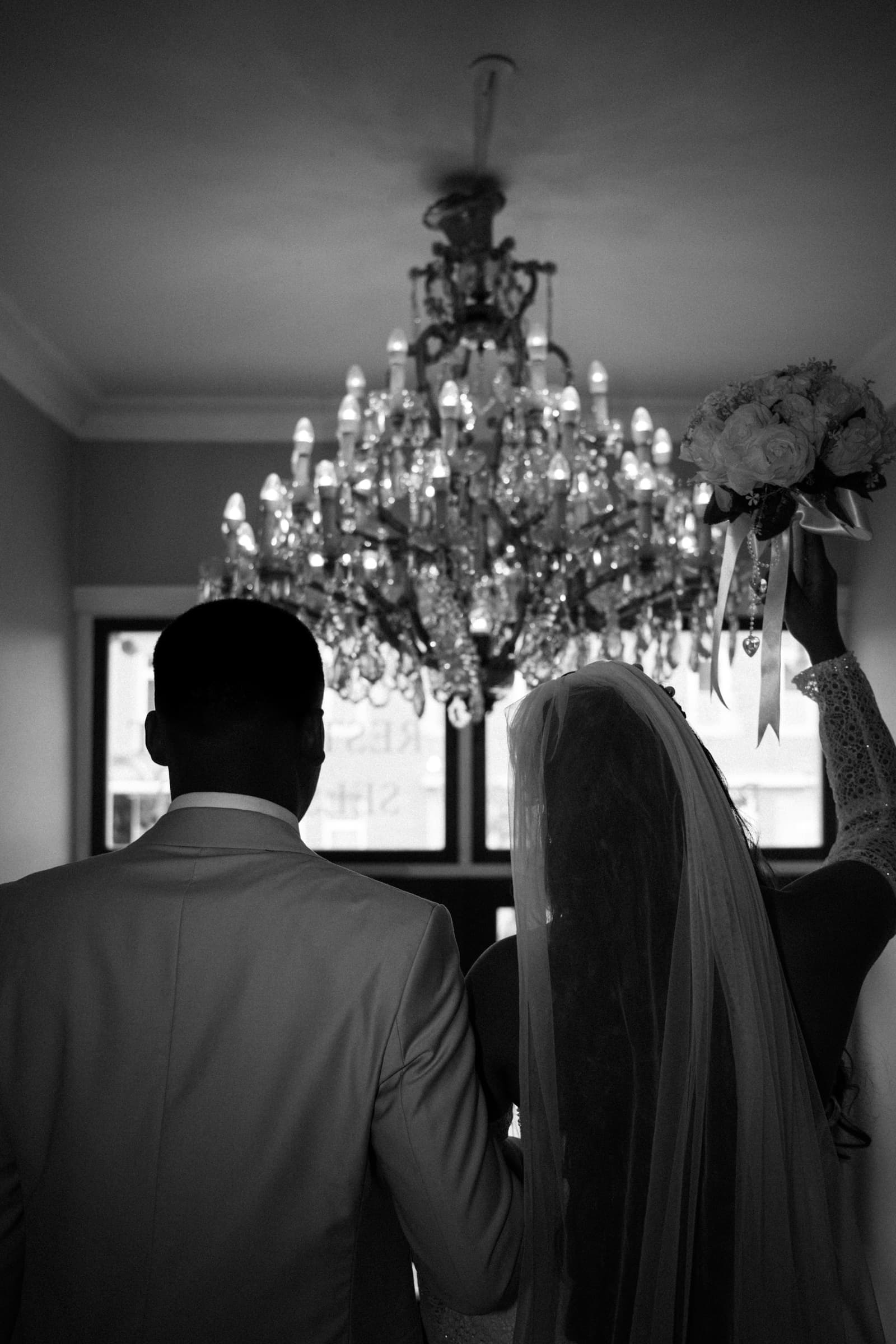 Couple from behind beneath a crystal chandelier, bouquet raised, black-and-white