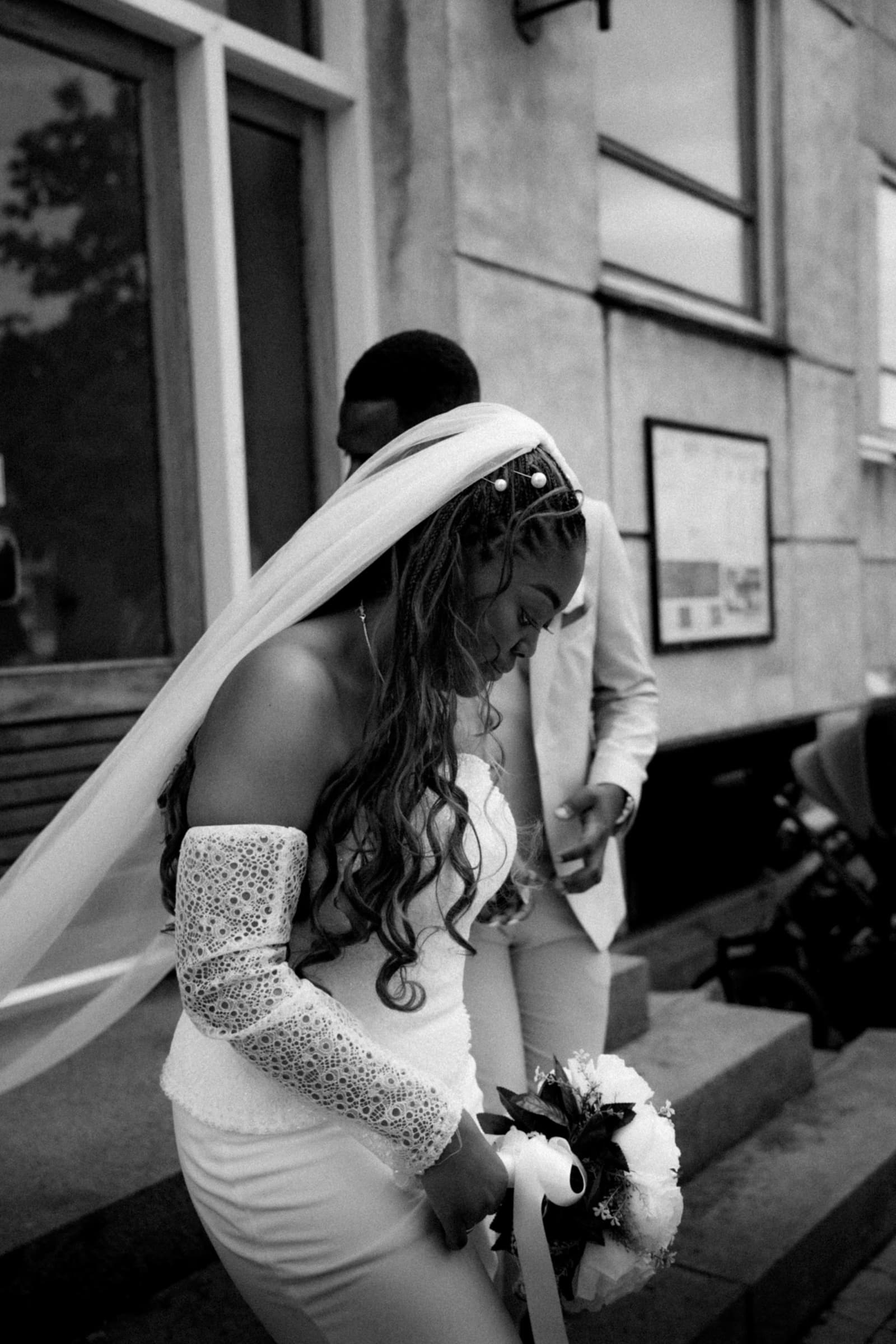 Bride in sharp focus adjusting her bouquet, groom soft behind her, black-and-white