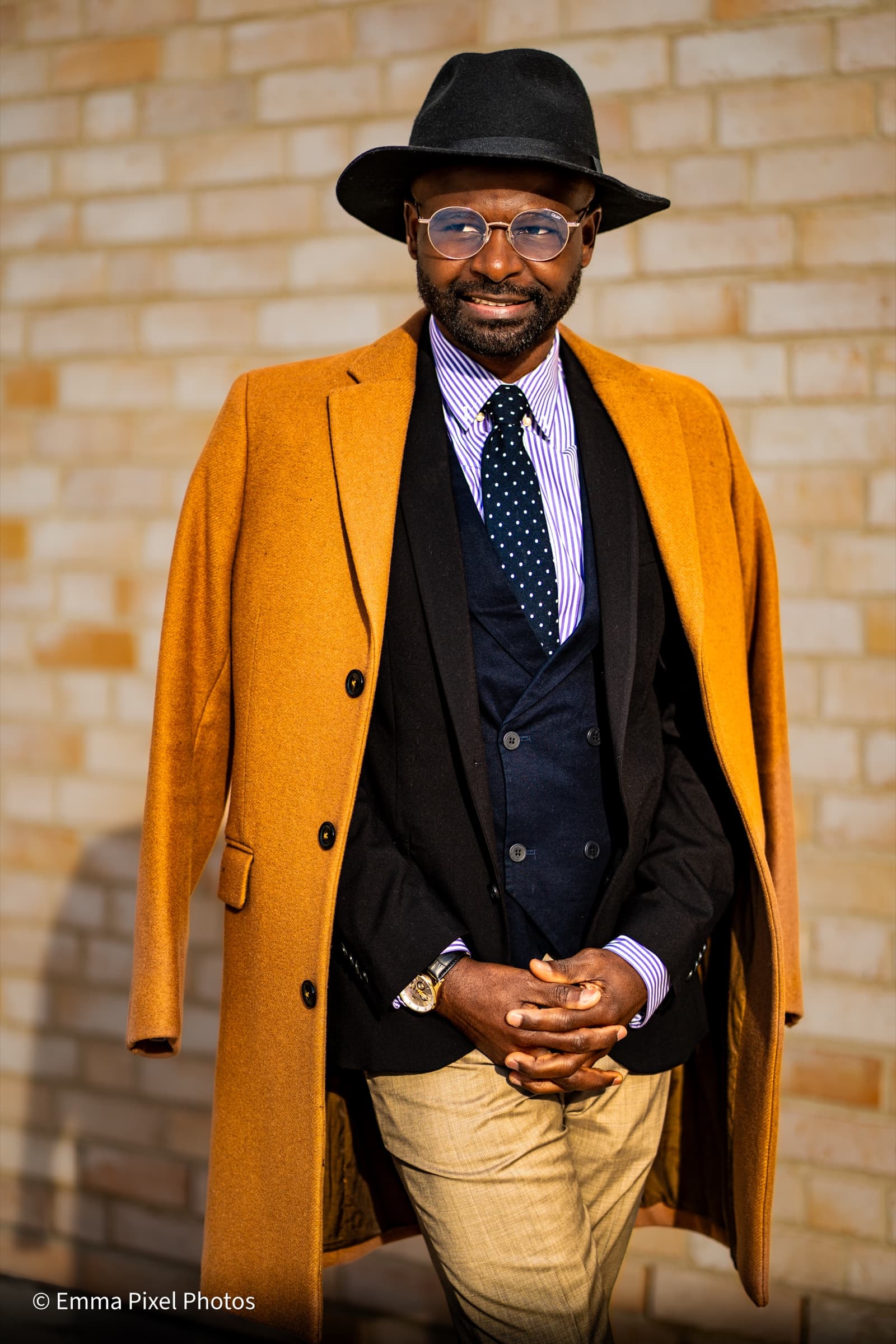 Hands clasped, mustard coat, brick wall portrait
