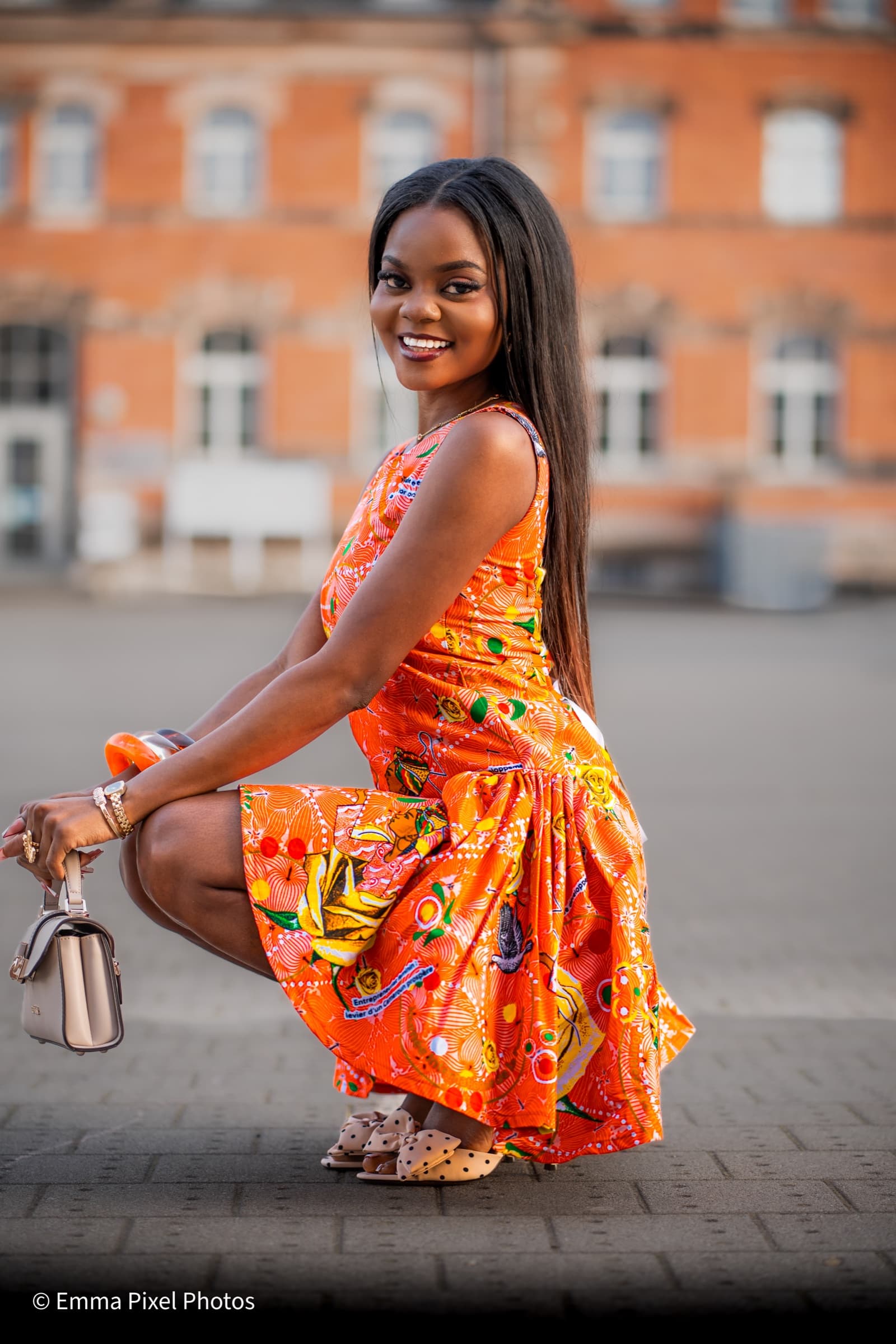 Orange African-print dress, crouching against red brick