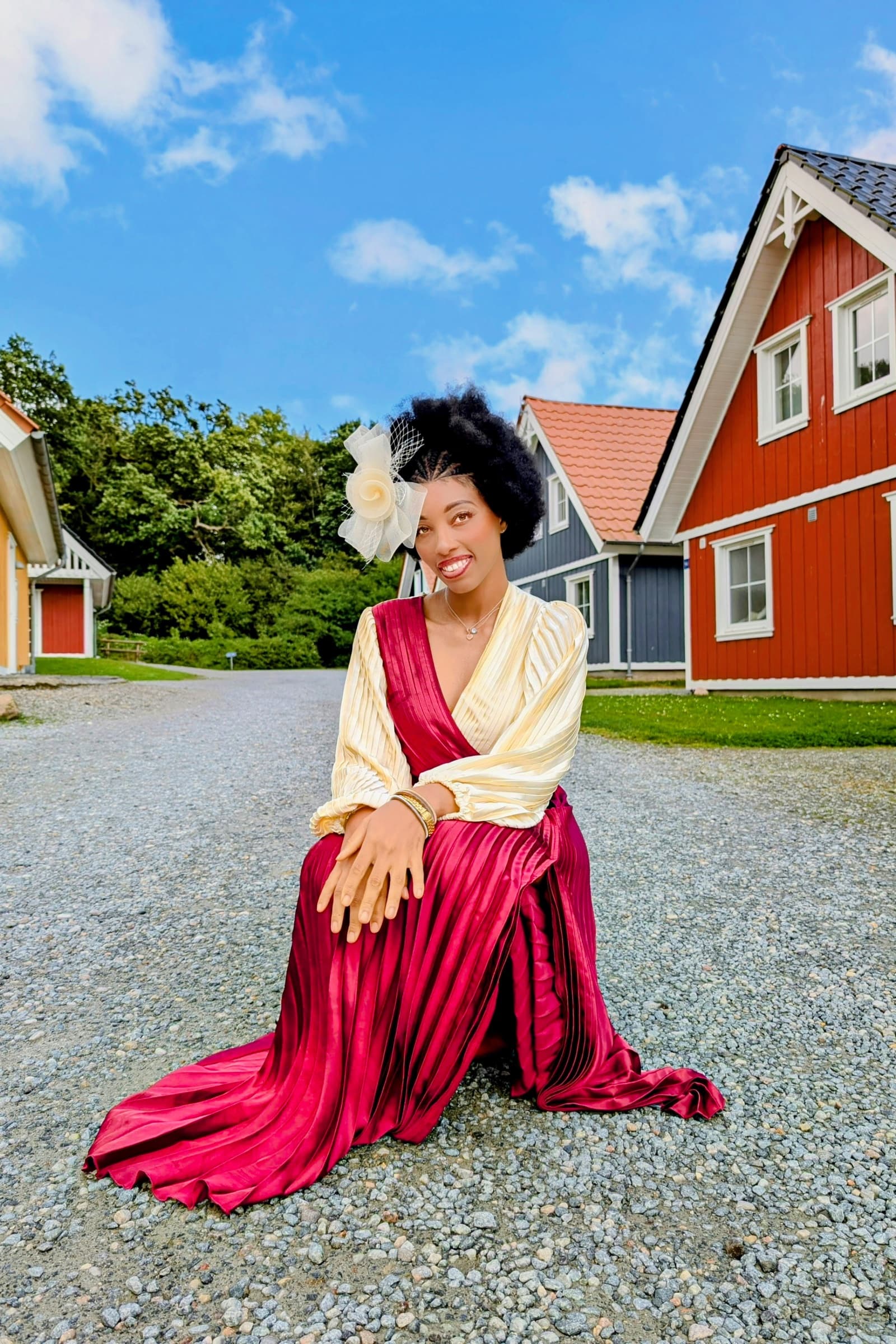 Burgundy gown and cream blouse, kneeling in front of red Nordic house
